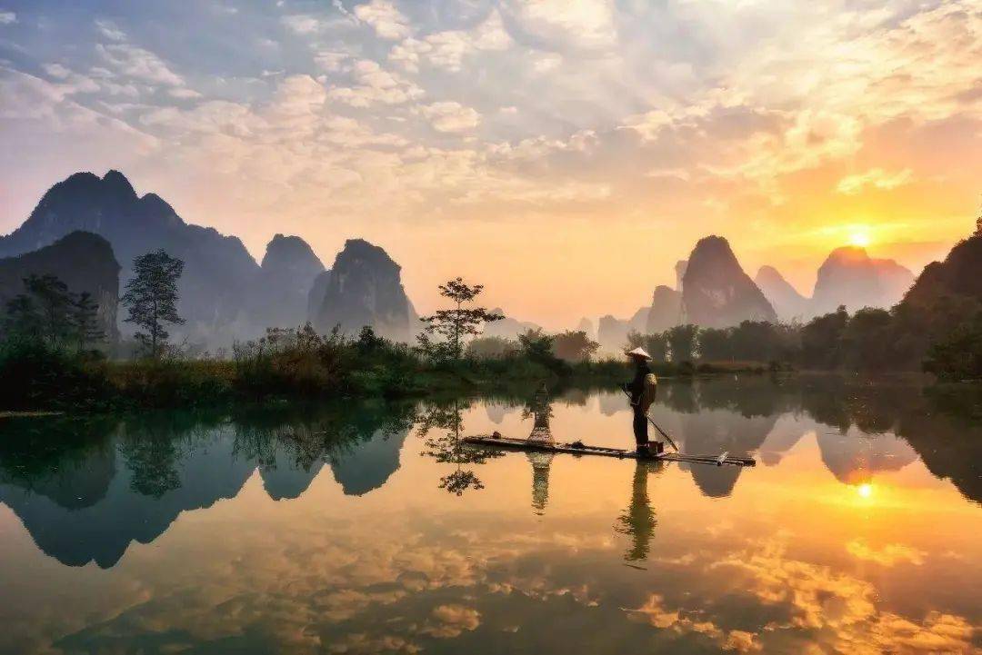 这是一张美丽的风景照片，展示了日出时刻，一个人在平静的河面上划着竹筏，远处是连绵的山峦和倒映在水中的倒影。